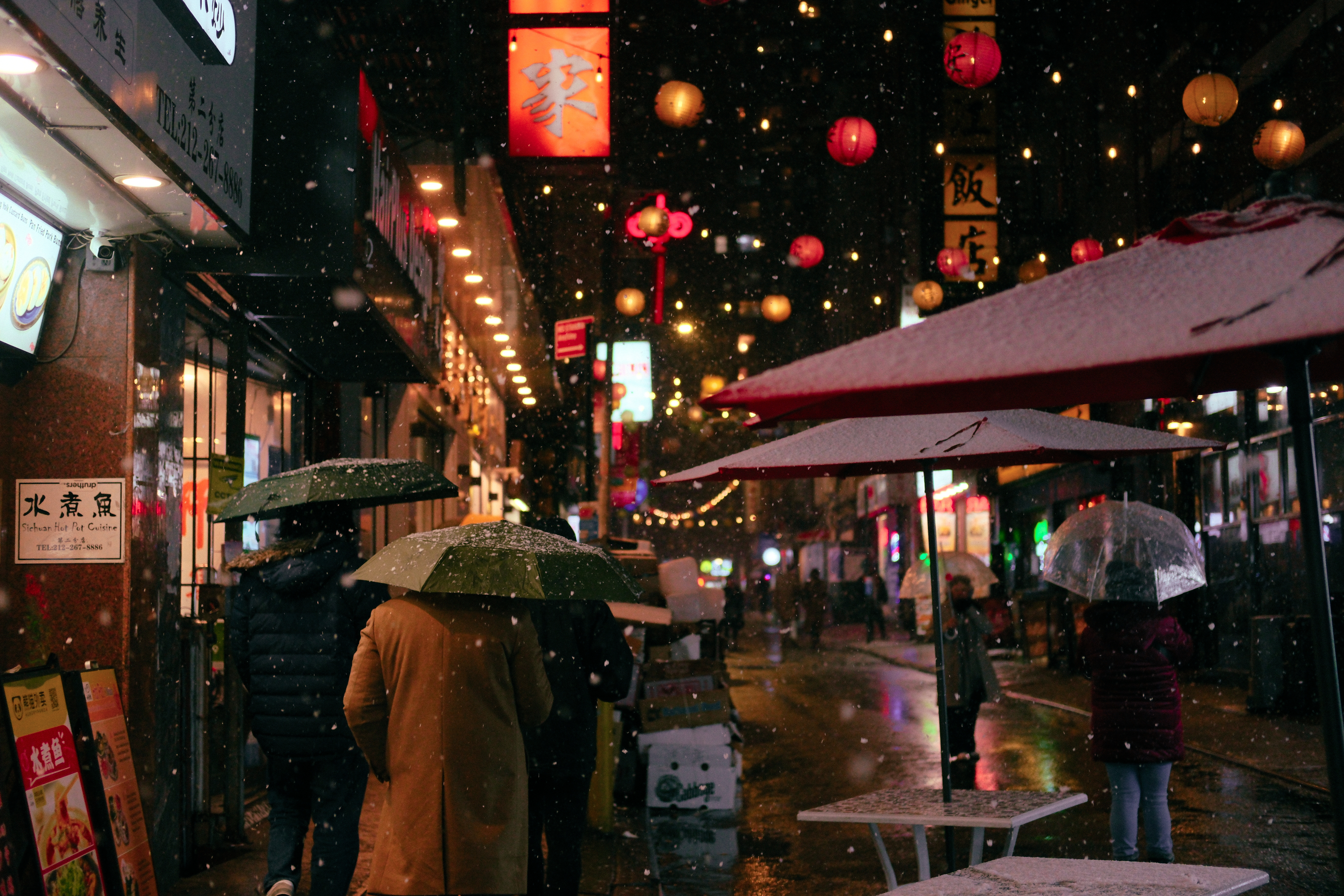 Snowy day in Chinatown at night. A few with umbrellas walk under red lanterns.